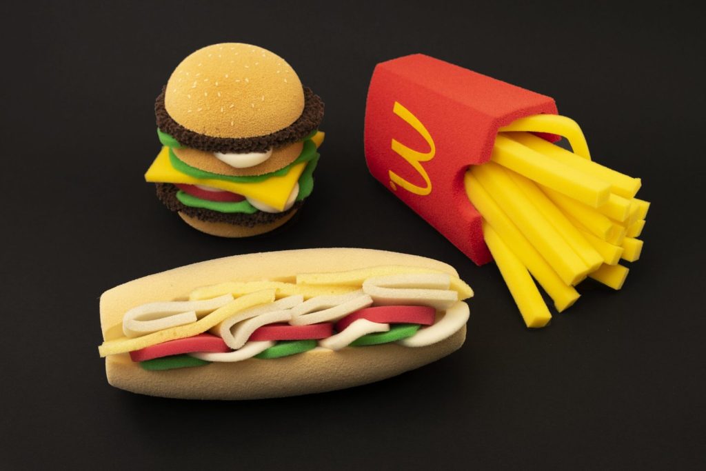 Custom-shaped foam microphone windshields styled as a burger, fries carton, and sandwich, showcasing bespoke 3D branded windscreens for broadcast and video production.