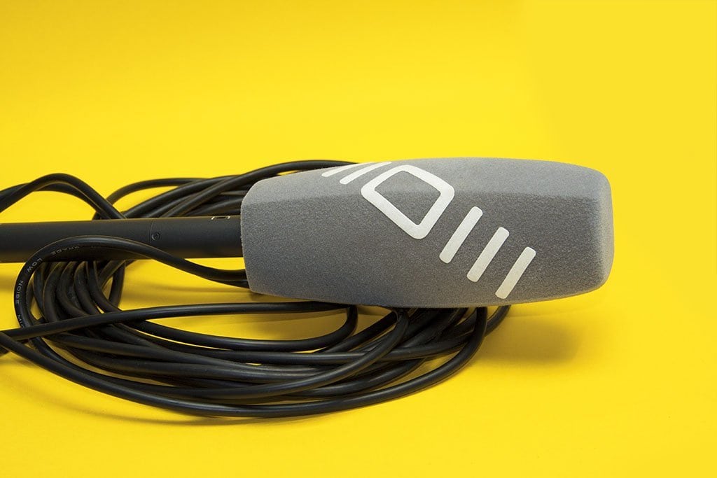 4-sided (square) grey foam microphone windscreen with a white logo, shown on a handheld microphone with coiled cable on a yellow background.