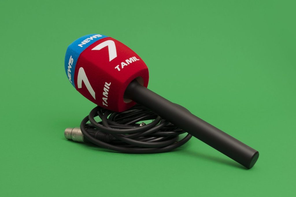 4-sided (square) red and blue foam microphone windscreen with “7 Tamil News” branding on a handheld microphone, shown with a coiled cable on a green background.