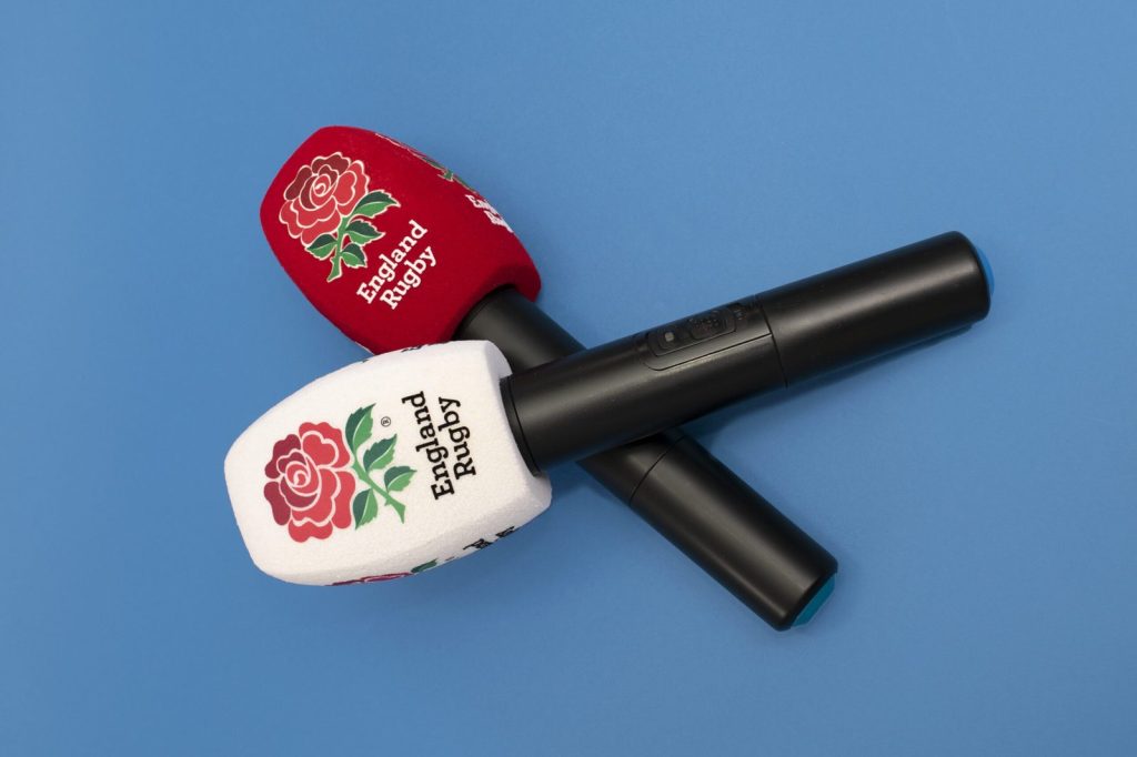 Two handheld microphones with foam windscreens branded “England Rugby” and a red rose logo, shown on a blue background (one red windscreen, one white).