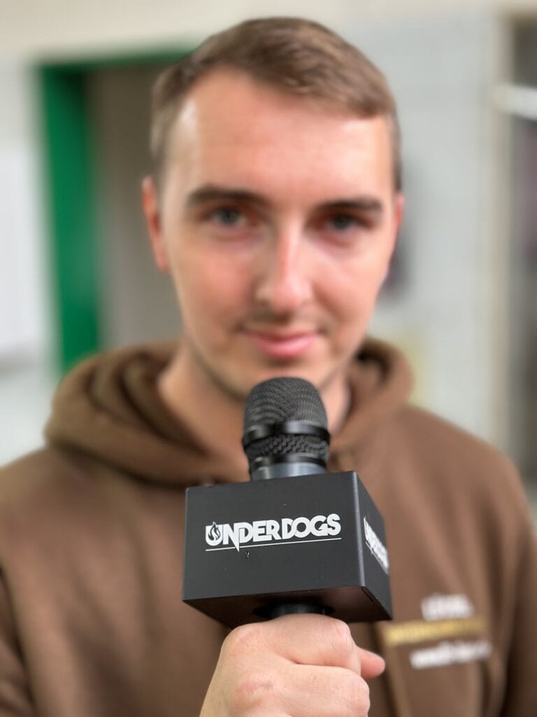 Black square microphone flag (mic cube) with white “UNDERDOGS” branding on a handheld interview microphone, broadcast and event reporting accessory.