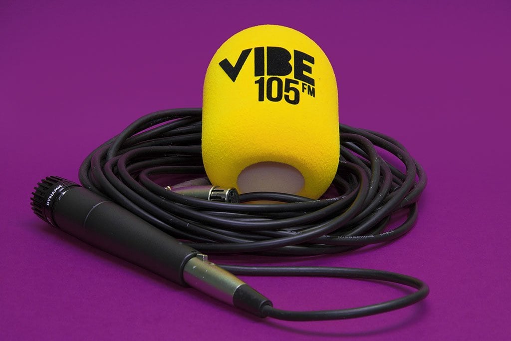 Yellow cylindrical foam microphone windscreen printed “VIBE 105 FM”, shown with a handheld microphone and coiled cable on a purple background.