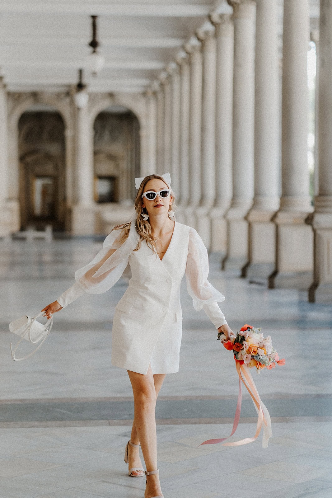 Eine Frau im eleganten weißen Kleid und mit Sonnenbrille schreitet selbstbewusst eine große Kolonnade hinunter, in der einen Hand einen bunten Blumenstrauß mit Bändern, die die Eleganz der Hochzeitsplanung Bayern widerspiegeln, in der anderen einen weißen Hut.