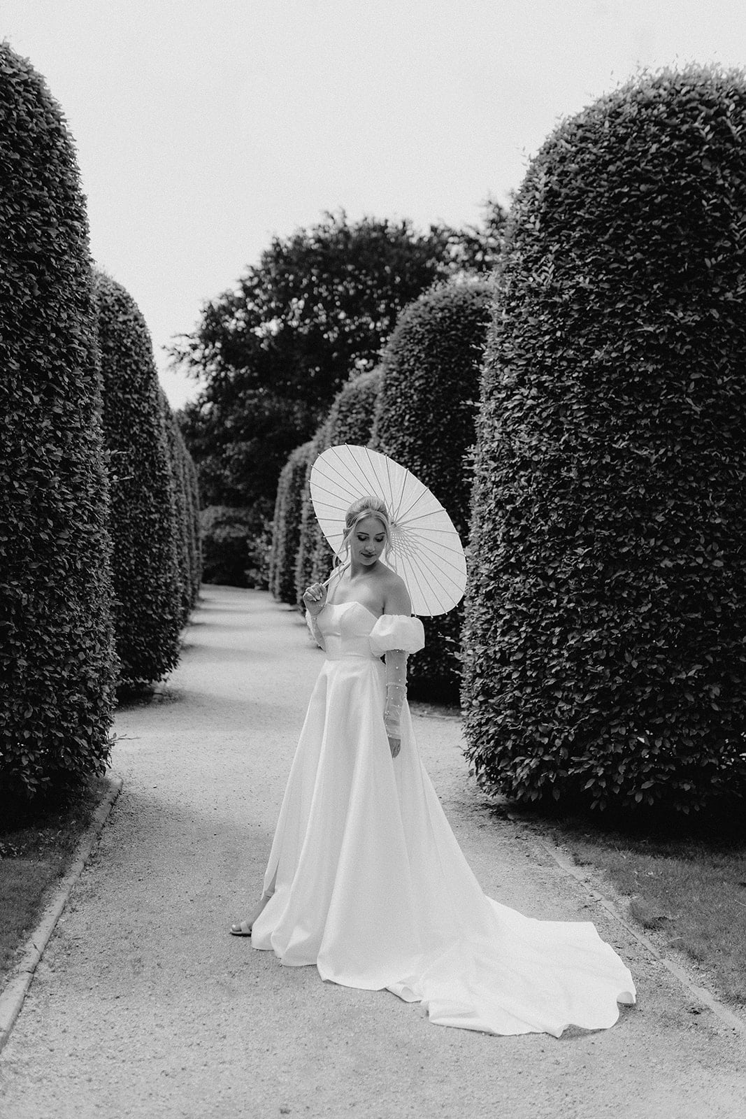 Eine Braut in einem langen, eleganten Kleid steht auf einem Gartenweg zwischen hohen, gemeißelten Hecken und hält einen Sonnenschirm. Dieses Schwarz-Weiß-Bild fängt die zeitlose Schönheit ein, die Hochzeitsplaner Nürnberg für jeden besonderen Tag bietet.