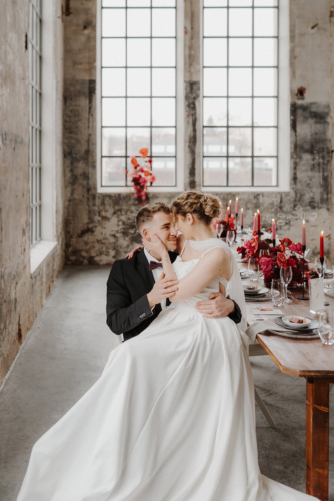 Ein Bräutigam im schwarzen Anzug hält seine Braut auf dem Schoß, beide lächeln und berühren sich an der Stirn in einem rustikalen, sonnendurchfluteten Raum mit hohen Fenstern und einem mit roten Kerzen und Blumen geschmückten Tisch - die perfekte Inspiration für Ihren Hochzeitsplaner Bayreuth.