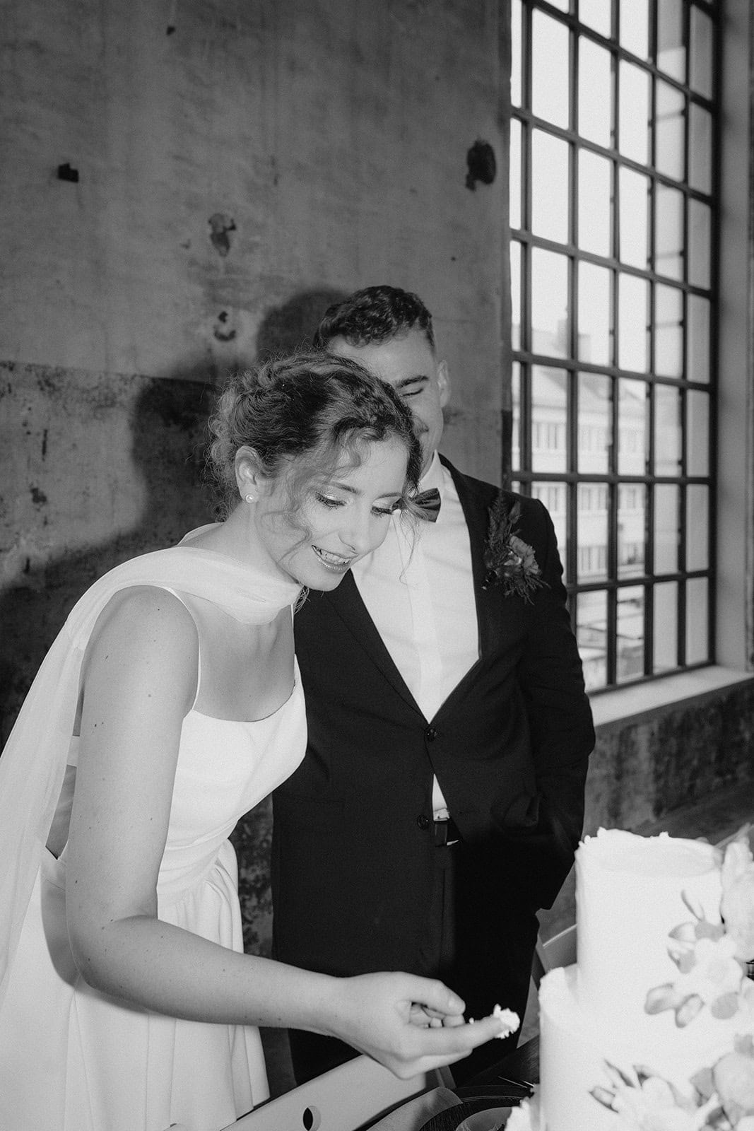 Eine lächelnde Braut in einem weißen Kleid schneidet eine Hochzeitstorte neben ihrem Bräutigam in einem Lokal im Industriestil mit großen Fenstern an. Die in Schwarz-Weiß aufgenommene Szene spiegelt den zeitlosen Touch eines Weddingplaner Bayern wider.