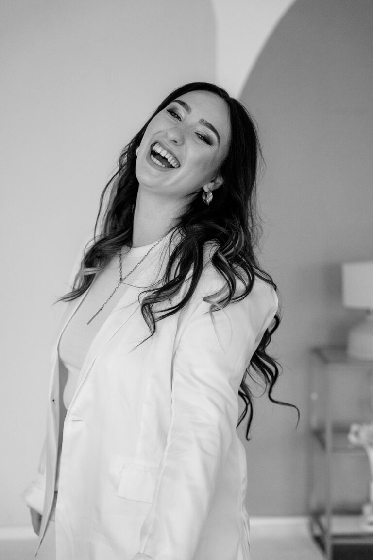 Eine Frau mit langen gewellten Haaren, die einen hellen Blazer und eine Halskette trägt, lächelt breit und lacht in einem sanft beleuchteten Raum. Das Schwarz-Weiß-Bild fängt perfekt die fröhliche Stimmung eines Hochzeitsplaners in Nürnberg ein.
