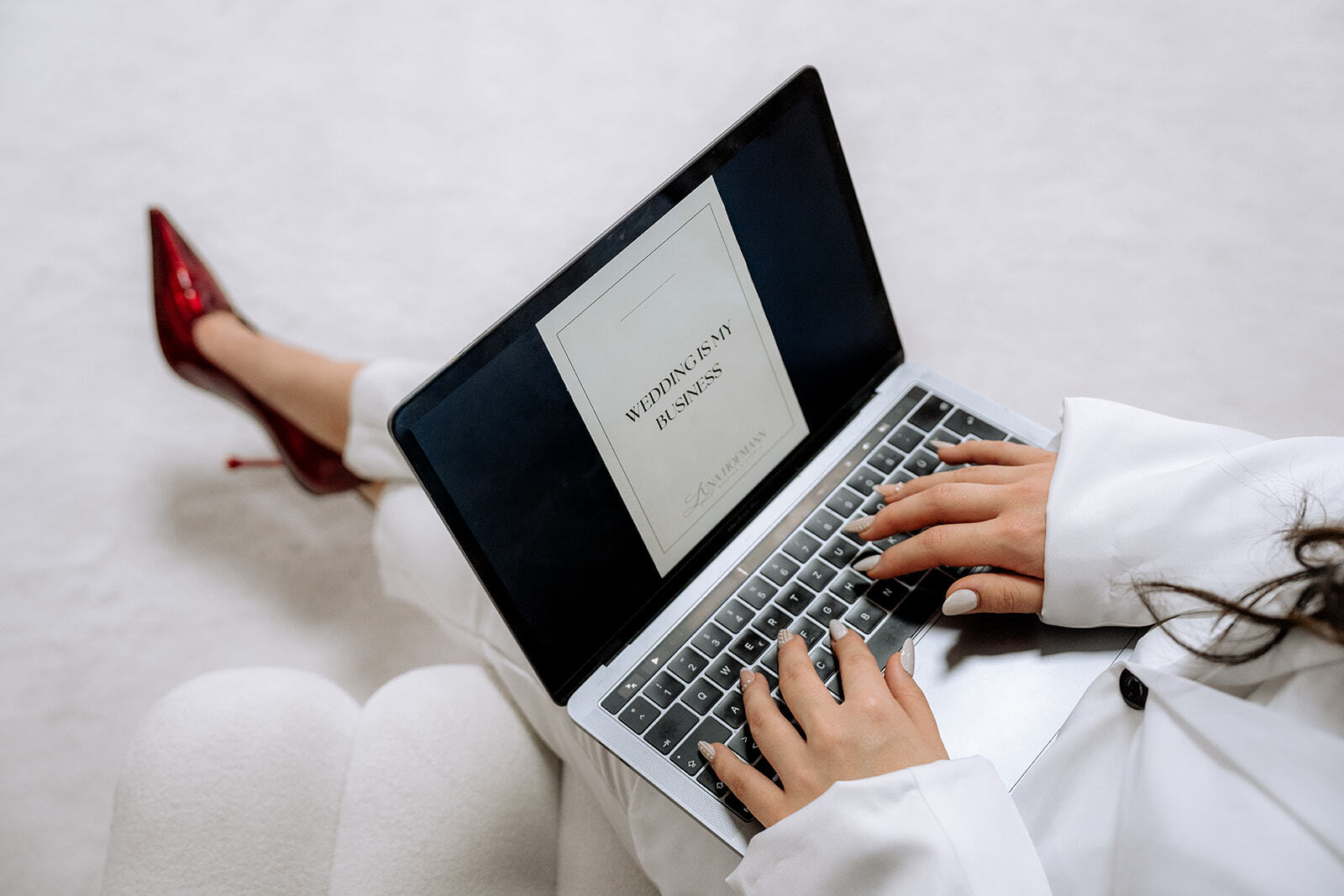 Eine Person in weißer Kleidung tippt auf einem Laptop, der eine Hochzeitseinladung anzeigt - nur ihre Hände und Beine in roten High Heels sind zu sehen. Die perfekte Inspiration für alle Hochzeitsplaner Bayern oder Hochzeitsplaner Nürnberg, die den großen Tag organisieren.