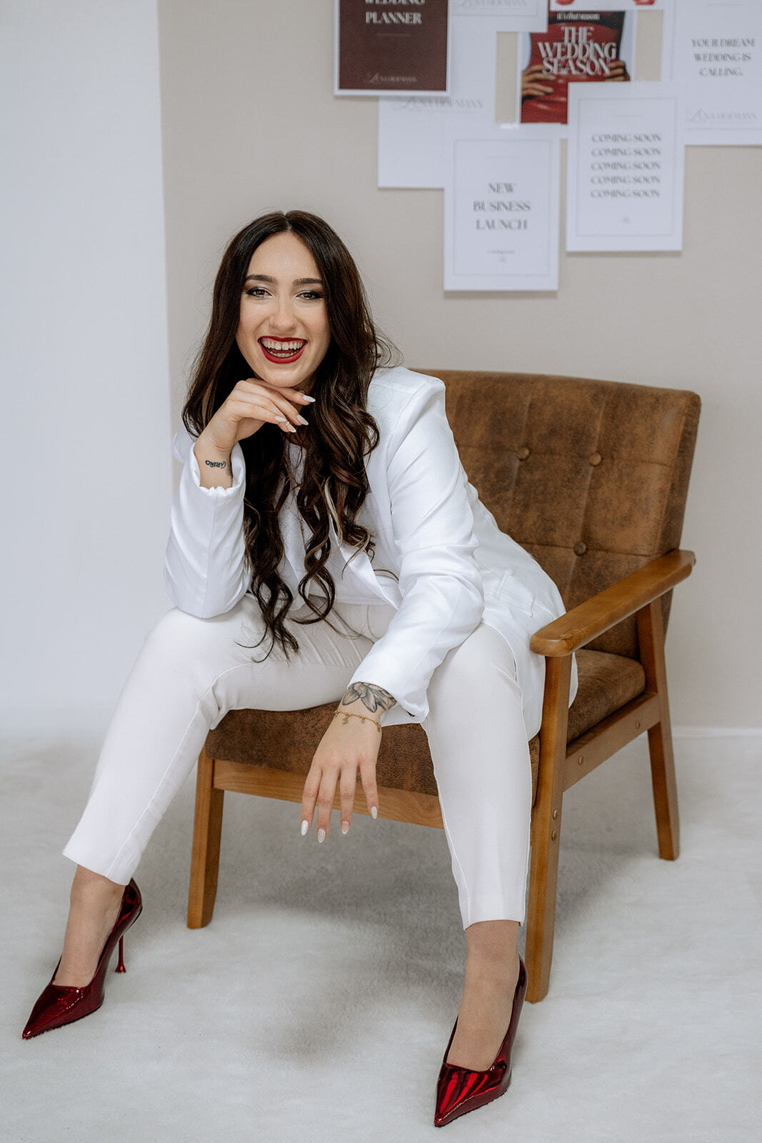 Eine Frau mit langen dunklen Haaren, weißem Anzug und roten High Heels, sitzt selbstbewusst auf einem braunen Stuhl, lächelt und stützt ihr Kinn auf die Hand. Hinter ihr stehen motivierende Zitate darüber, ein Top-Hochzeitsplaner Bayern zu sein.