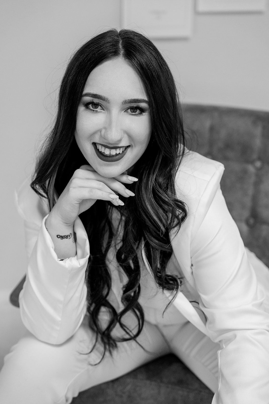Eine Frau mit langen dunklen Haaren und einem strahlenden Lächeln, gekleidet in einen weißen Anzug, sitzt auf einem Stuhl und lehnt sich vor, das Kinn auf ihre Hand gestützt. Dieses Schwarz-Weiß-Bild fängt die Eleganz eines professionellen Hochzeitsplaners Bayreuth ein.