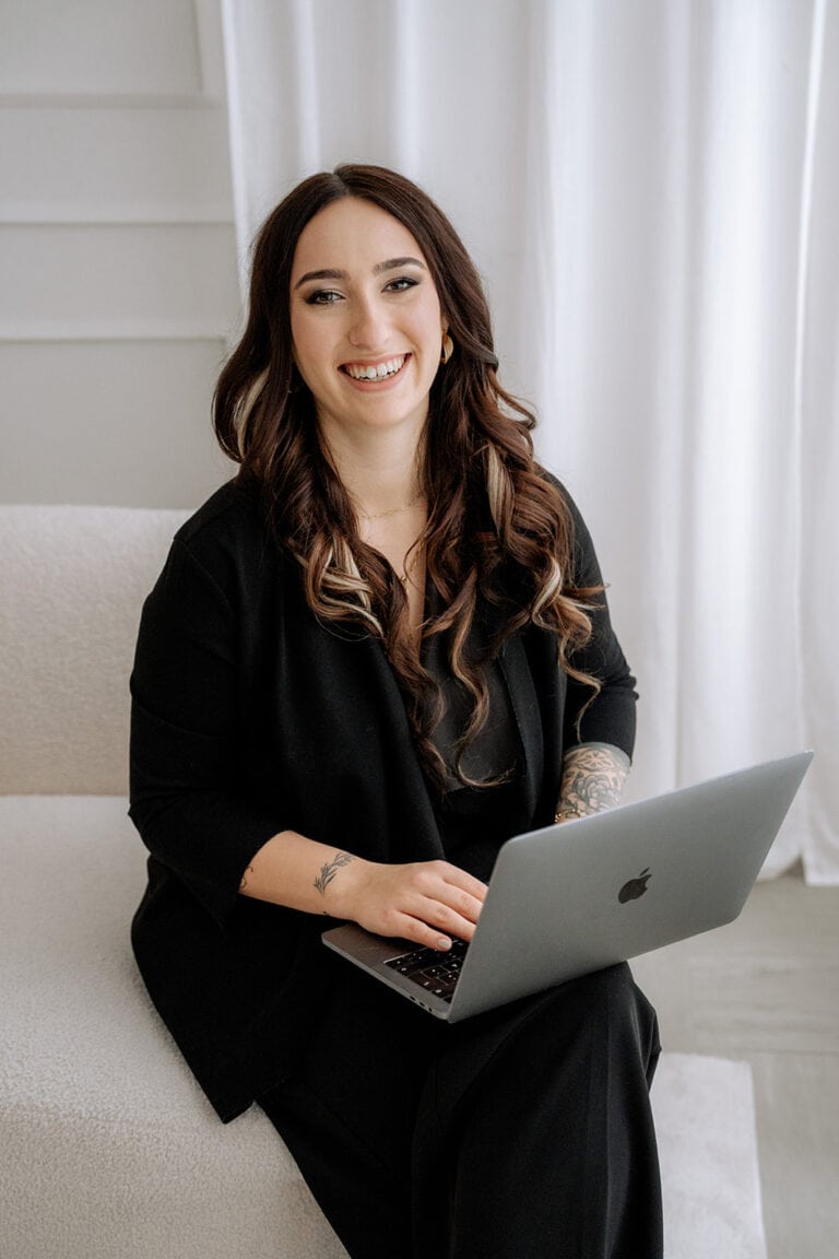 Eine Frau mit langen braunen Haaren und Tattoos, schwarz gekleidet, sitzt lächelnd auf einem hellen Sofa und arbeitet an einem silbernen Laptop - beschäftigt mit Hochzeitsplanung Bayern. Im Hintergrund sind weiße Vorhänge und eine neutrale Wand zu sehen.