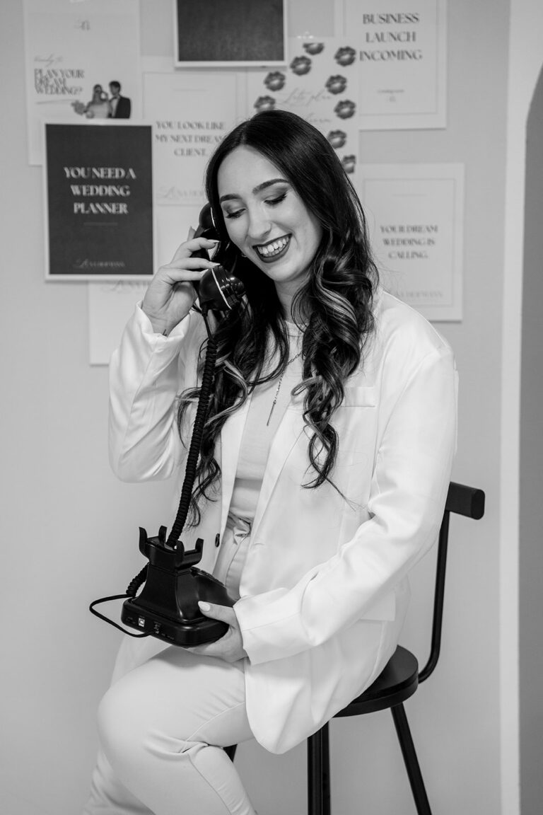 Eine lächelnde Frau mit langen Haaren in einem hellen Anzug sitzt auf einem Hocker und telefoniert mit einem alten Telefon. Hinter ihr sind Motivationsplakate zu sehen - die perfekte Inspiration für jeden Hochzeitsplaner Nürnberg. Das Foto ist in schwarz-weiß gehalten.