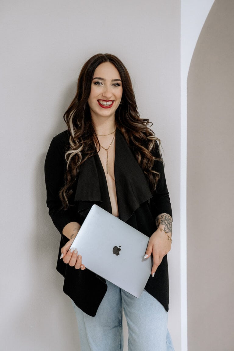Eine Frau mit langen braunen Haaren und hellen Strähnchen lächelt, während sie vor einer hellen Wand steht, eine schwarze Jacke und helle Jeans trägt und einen geschlossenen silbernen Laptop in der Hand hält - perfekt für alle Hochzeitsplaner Bayern, die bereit sind, ihre Traumhochzeit zu planen.