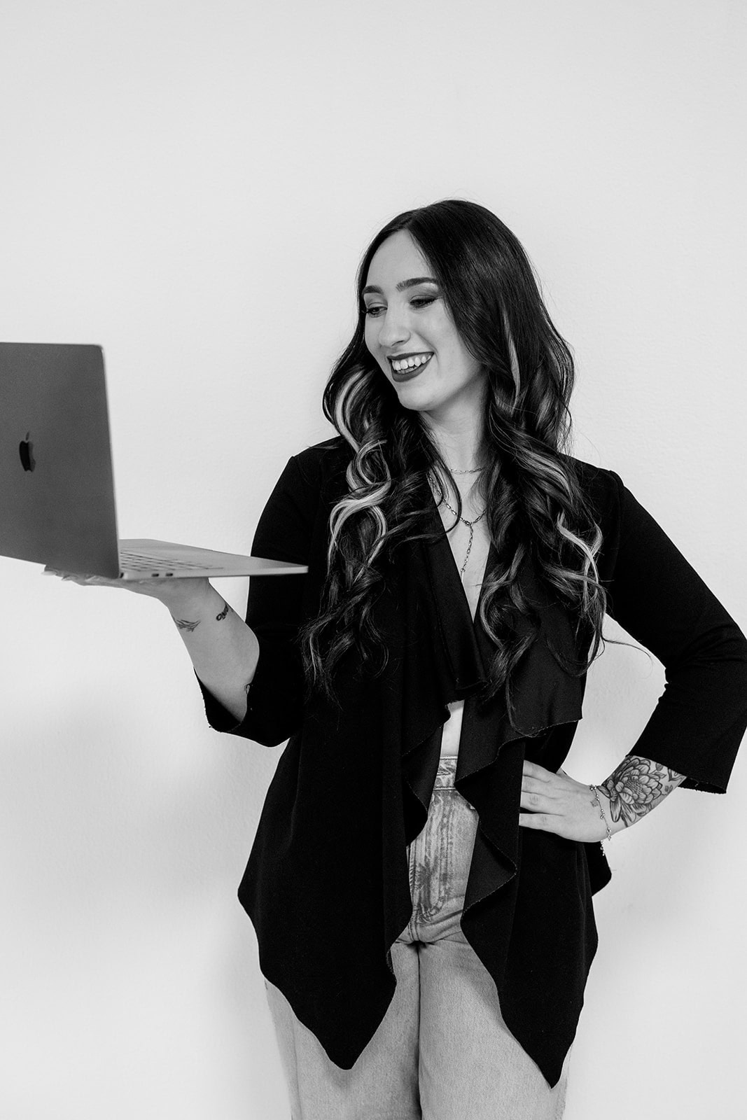 Eine Frau mit langen gewellten Haaren und Tattoos lächelt, während sie einen geöffneten Laptop in der Hand hält und den kreativen Geist eines Hochzeitsplaners Nürnberg verkörpert, während sie selbstbewusst vor einem schlichten weißen Hintergrund steht.