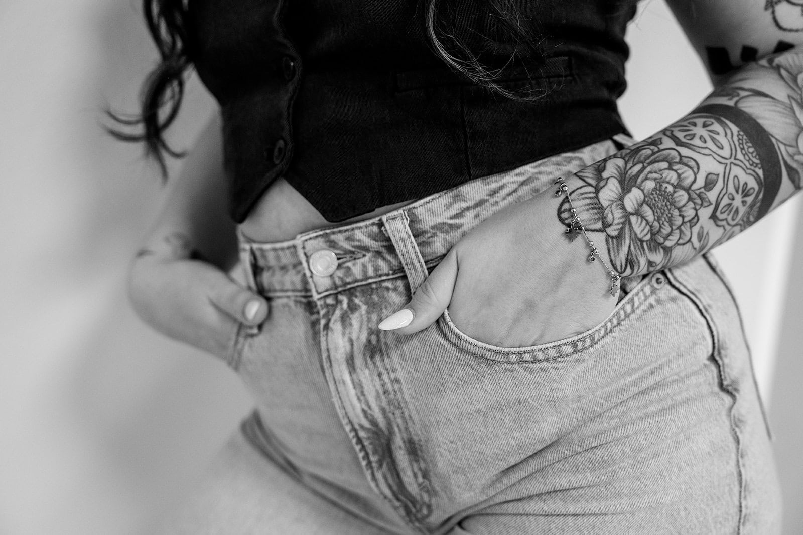 Eine Person in High-Waisted-Jeans und einem ärmellosen Top, die Hände in den Taschen, zeigt ein detailliertes Blumentattoo. Dieses in Schwarz-Weiß aufgenommene Bild spiegelt den schicken Stil wider, den man oft bei Veranstaltungen der Hochzeitsplanung Bayern sieht.