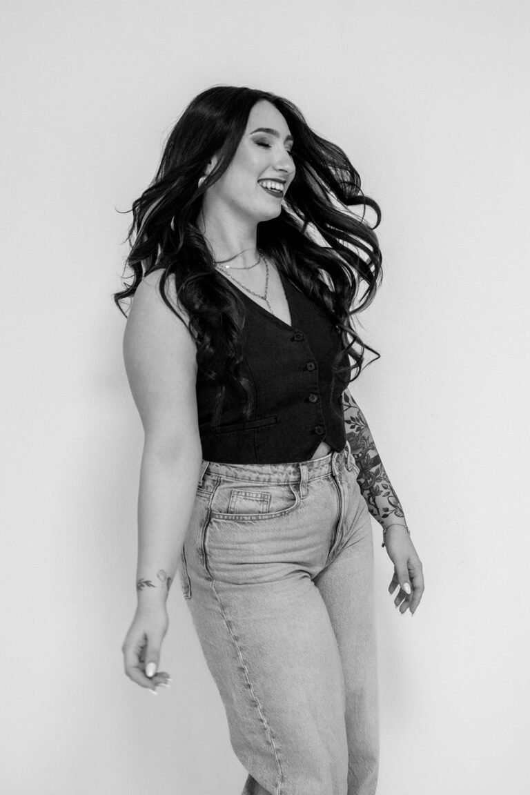 Eine Frau mit langem, gewelltem Haar lächelt und schaut zur Seite, während sie vor einem schlichten Hintergrund steht. Sie trägt ein ärmelloses schwarzes Oberteil, eine Jeans mit hoher Taille und hat Tattoos auf dem linken Arm - die perfekte Inspiration für einen modernen Weddingplaner Bayern. Das Foto ist in schwarz-weiß gehalten.