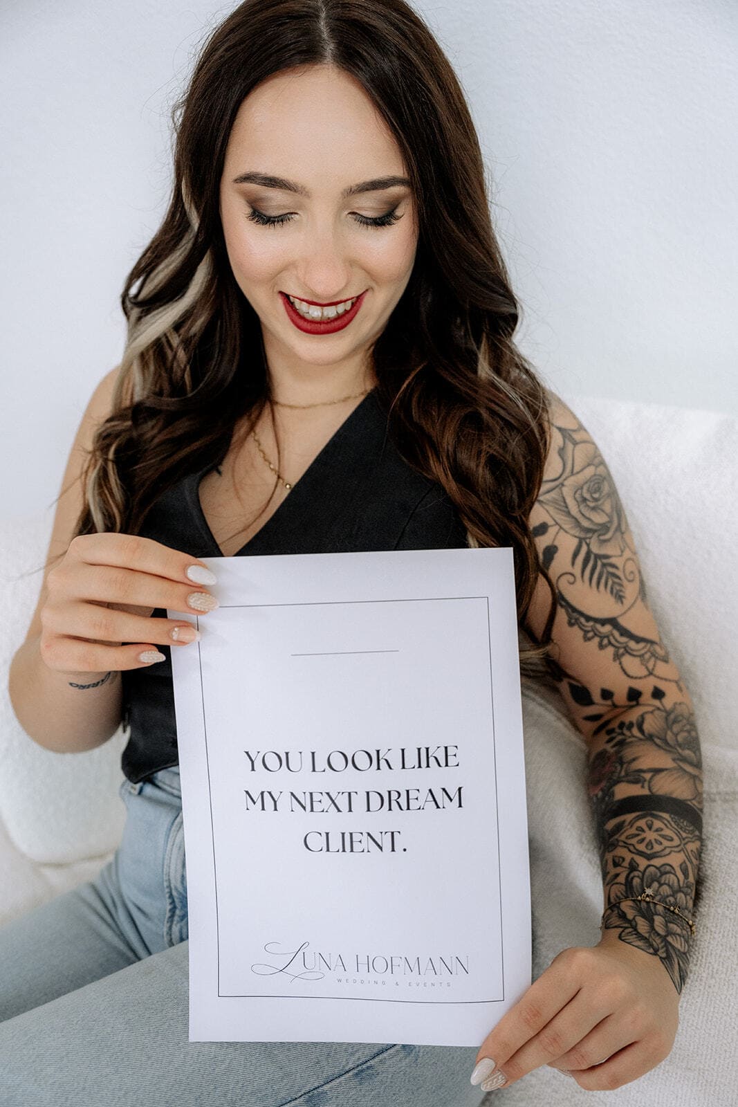 Eine lächelnde Frau mit langen braunen Haaren und einem tätowierten Arm, bekannt als Luna Hofmann, hält ein Schild mit der Aufschrift "Sie sehen aus wie mein nächster Traumkunde". Sie sitzt auf einer hellen Fläche - Ihre Expertin für Hochzeitsplanung Bayern.