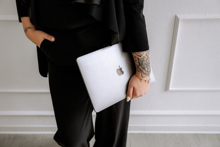 Eine Person in schwarzer Kleidung hält ein geschlossenes silbernes MacBook, eine Hand in der Tasche. Mit Tattoos auf dem Arm steht sie vor einer weißen Wand - und fängt damit perfekt die kreative Stimmung eines modernen Hochzeitsplaners in Bayreuth ein.