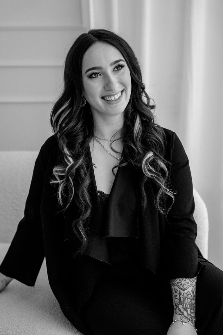 Schwarz-Weiß-Foto einer Frau mit langem, gewelltem Haar, die auf einem Stuhl sitzt, lächelt und leicht zur Seite schaut. Sie trägt ein dunkles Outfit, zeigt ein Tattoo auf ihrem linken Unterarm - ein elegantes Porträt eines Hochzeitsplaners Bayern in Aktion.