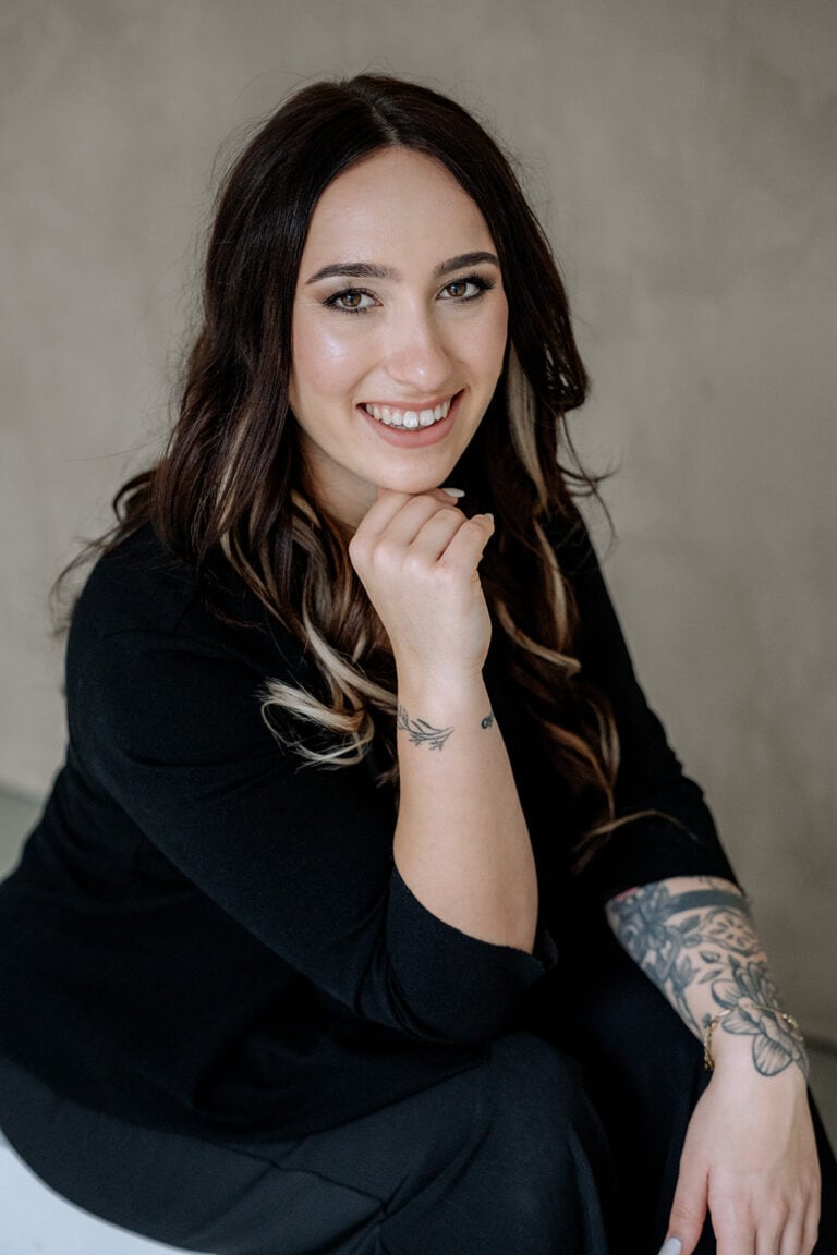 Eine Frau mit langen braunen Haaren und Tätowierungen auf dem rechten Arm, vielleicht ein Hochzeitsplaner Bayern, lächelt in die Kamera und stützt ihr Kinn auf ihre Hand. Sie trägt ein schwarzes Oberteil und sitzt vor einem neutralen, hellen Hintergrund.