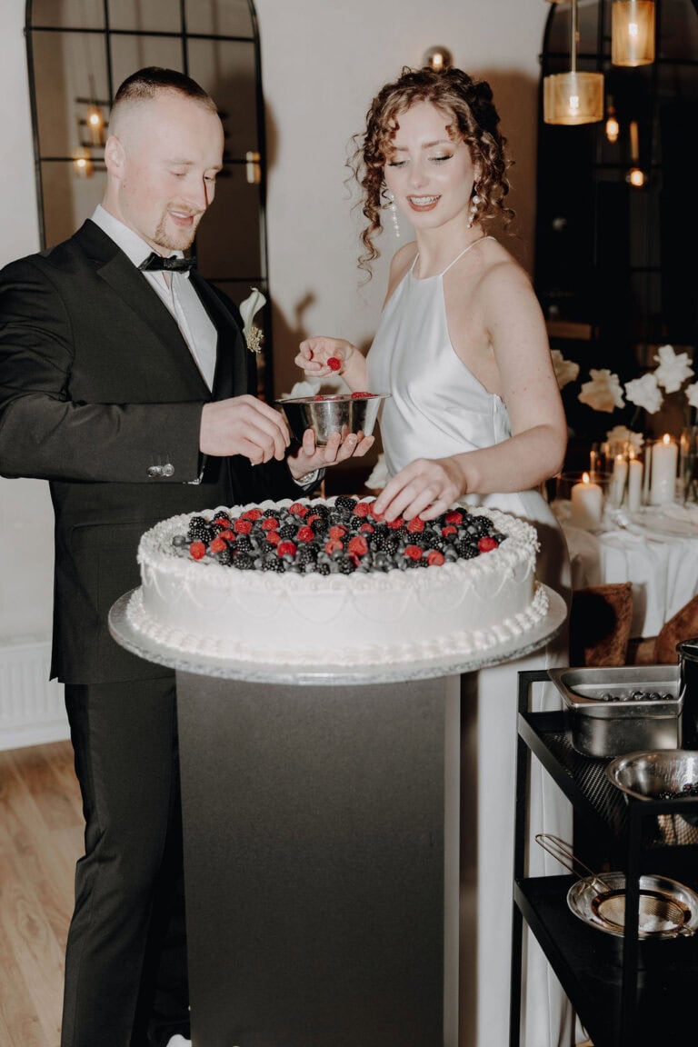 Ein Mann in einem schwarzen Anzug und eine Frau in einem weißen Kleid, begleitet von einem Hochzeitsplaner Bayern, dekorieren eine große Torte mit Beeren bei einer eleganten, kerzenbeleuchteten Veranstaltung, lächelnd und konzentriert als Teil ihrer nahtlosen Hochzeitsfeier.