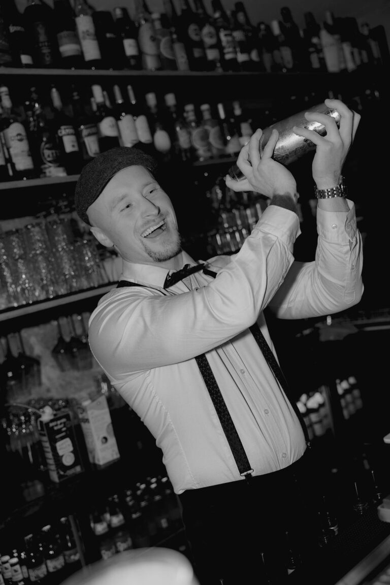 Ein Barkeeper in einem weißen Hemd, Hosenträgern und einer flachen Kappe lächelt, während er einen Cocktailshaker hinter einer mit Flaschen und Gläsern bestückten Bar schüttelt. Dieses stilvolle Schwarz-Weiß-Bild ist perfekt für jedes Hochzeitsplaner Bayern oder Weddingplaner Portfolio.