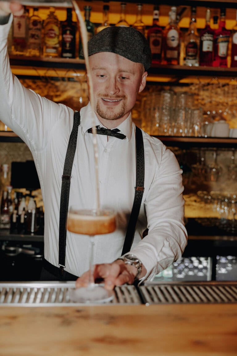 Ein Barkeeper in weißem Hemd, schwarzen Hosenträgern und Mütze schenkt an einer Bar einen Drink in ein Cocktailglas ein - die perfekte Inspiration für jeden Weddingplaner oder Hochzeitsplaner Bayreuth, der auf der Suche nach stilvollen Veranstaltungsdetails ist. Hinter ihm reihen sich Flaschen und Gläser in den Regalen.