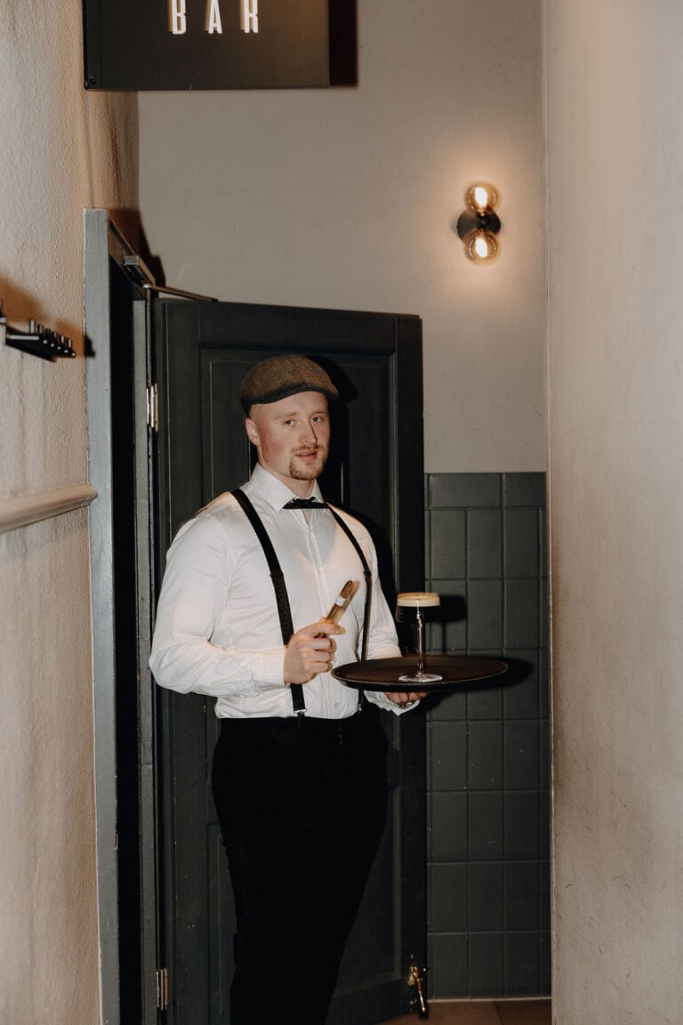Ein Kellner in weißem Hemd, schwarzen Hosenträgern und flacher Mütze steht im Flur und hält ein Tablett mit einem Glas und einem Flaschenöffner in der Hand - eine elegante Note, die man oft bei Veranstaltungen sieht, die von einem Hochzeitsplaner Nürnberg oder Weddingplaner Bayern organisiert werden.
