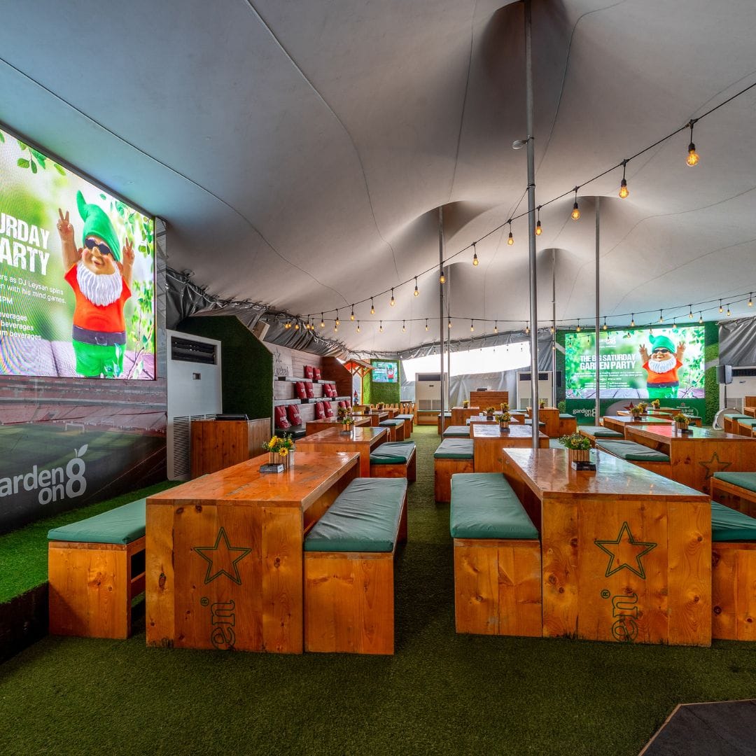 Garden on 8 outdoor seating with wooden tables, green turf, and festive decor under a canopy for a vibrant experience.