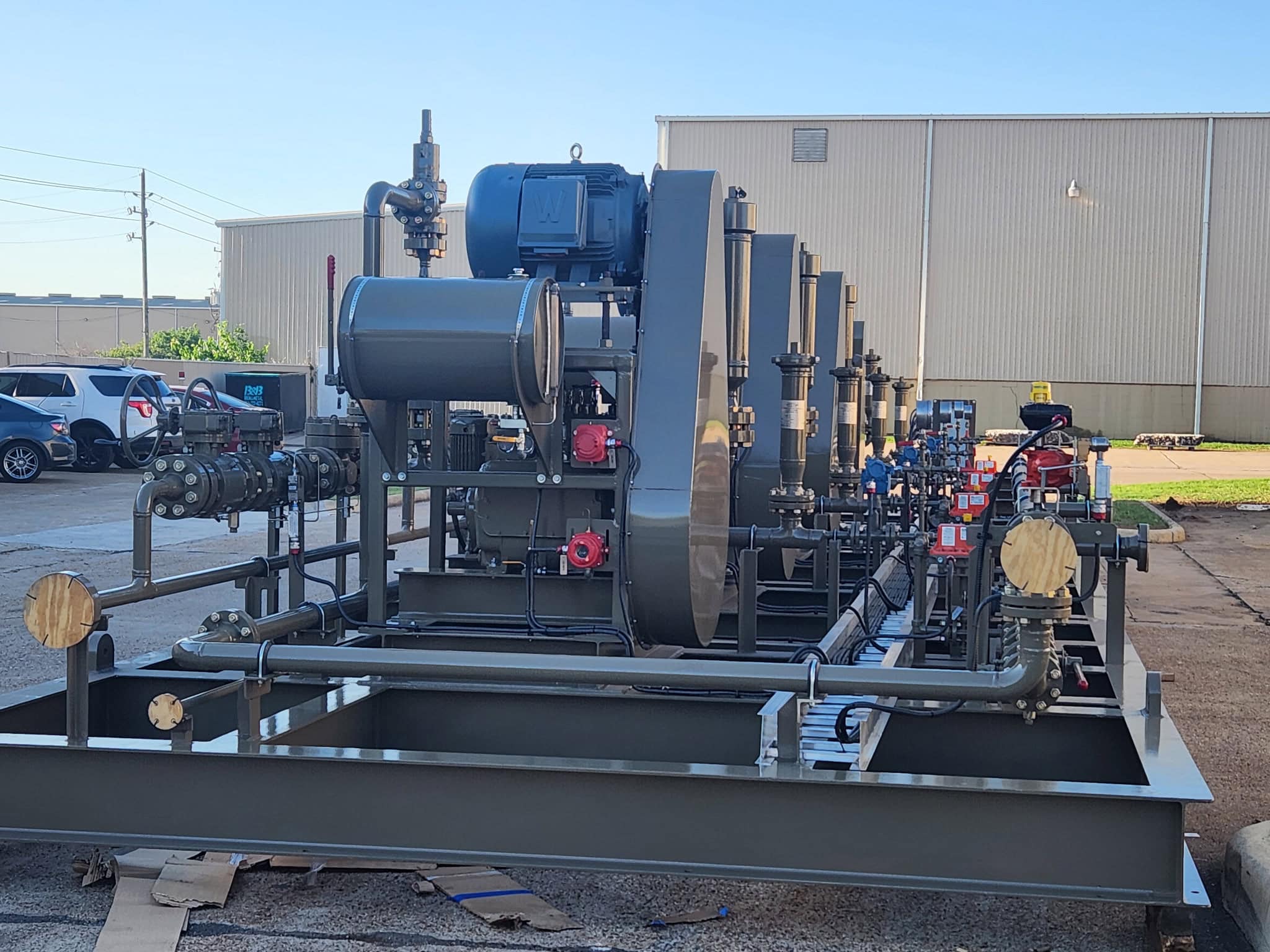 Large process pump skid with a blue motor, gray piping, valves and gauges mounted on a fabricated steel skid base outside a warehouse