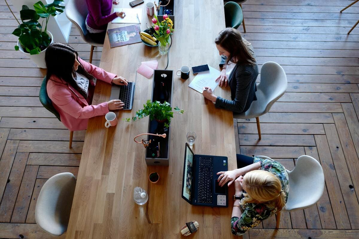 A group of professionals work together at a long wooden table in a modern coworking space with a herringbone-patterned wooden floor. The table is adorned with laptops, notebooks, a vase of pink flowers, bananas, a small potted plant, and various personal itens like mugs and water bottles. A large potted plant sits in the corner, and the overall atmosphere is bright and collaborative.