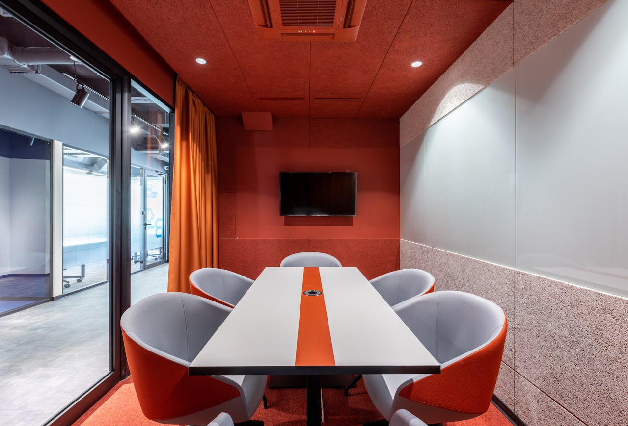 A modern Office Evolution meeting room features a long white table with an orange stripe down the center, surrounded by six gray chairs with orange accents. The room has a red acoustic ceiling and walls, with one wall displaying a mounted flat-screen TV. Orange curtains hang on one side, and large glass windows on the other reveal an adjacent office space with visible furniture and decor. The overall design is sleek and professional, with a warm, collaborative vibe.