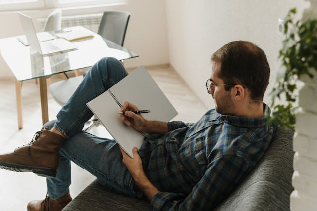 A person in a plaid shirt, jeans, and brown boots is seated on a gray couch in a modern Office Evolution coworking space, writing in a notebook. The background includes a glass table with a laptop and papers, an office chair, and a potted plant near a window, creating a relaxed and productive atmosphere. details on Office Evolution coworking space benefits