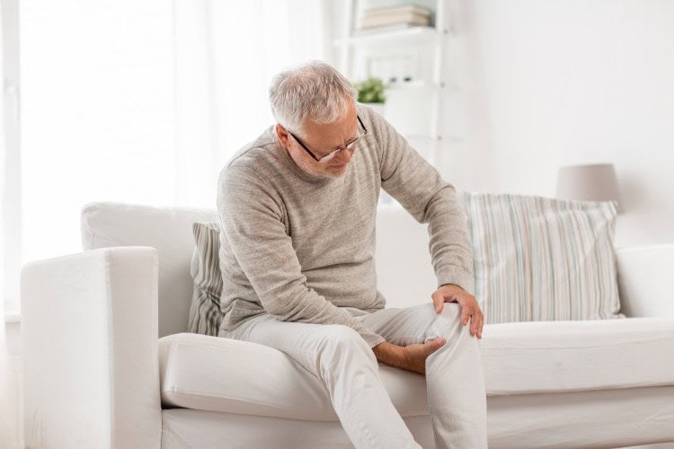 Senior man sitting with visible knee pain discomfort - One Body LDN Senior man sitting with visible knee pain discomfort