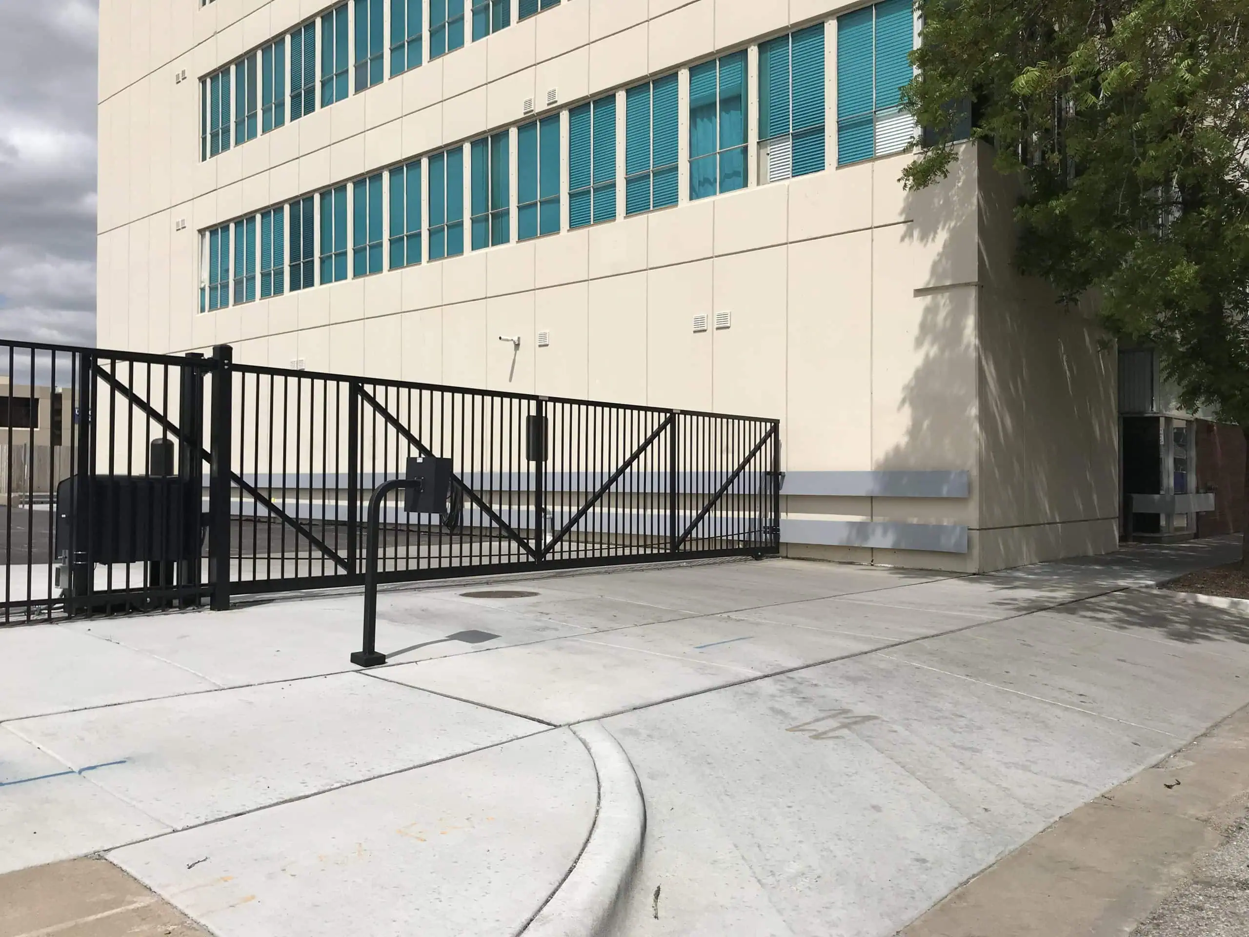 Commercial vehicular slide gate with black powder-coated steel vertical picket fencing at modern office building entrance. Industrial security sliding gate system featuring horizontal track design, concrete driveway access, and professional gate operator mounting hardware. Commercial property access control with heavy-duty steel slide gate construction, vertical bar infill panels, and automated sliding gate infrastructure at multi-story office complex with teal window accents and concrete facade.