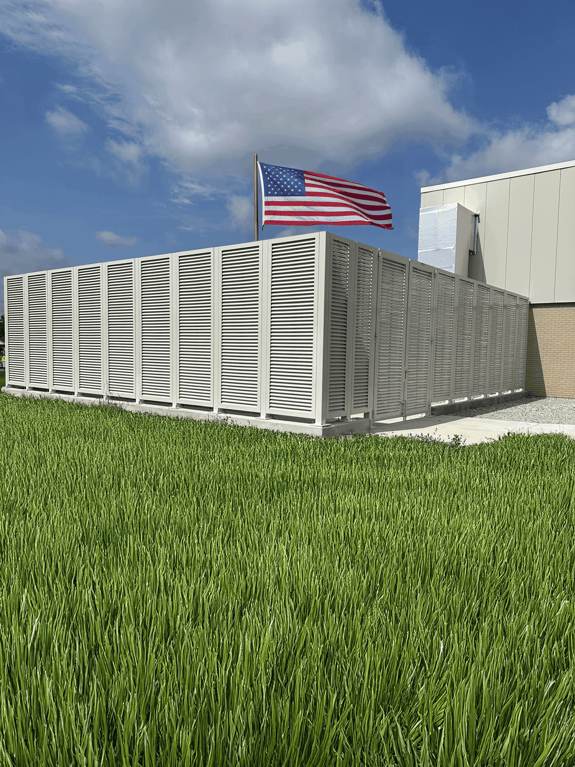 White PalmSHIELD ground screen enclosure protecting corporate building's mechanical equipment next to a field of luscious green freshly cut grass and American flag blowing in the wind directly behind the mechanical equipment screen enclosure