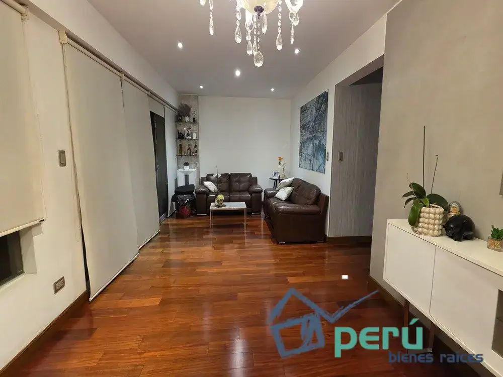Living room with two brown leather sofas, glass coffee table, chandelier, and wooden floor; white walls and decorative plant display on a sideboard.