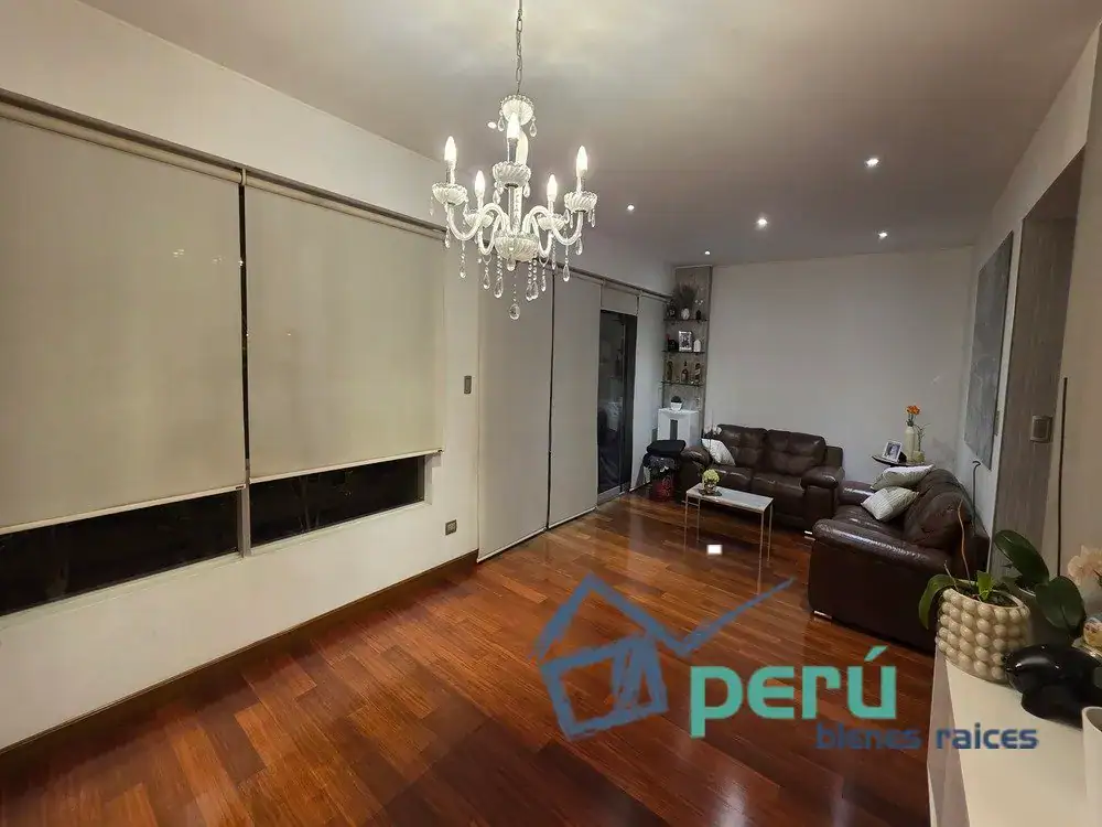 Bright living room with dark wood floor, a crystal chandelier, and large white blinds along one wall; modern sliding doors and a brown leather sectional seating area in the corner.