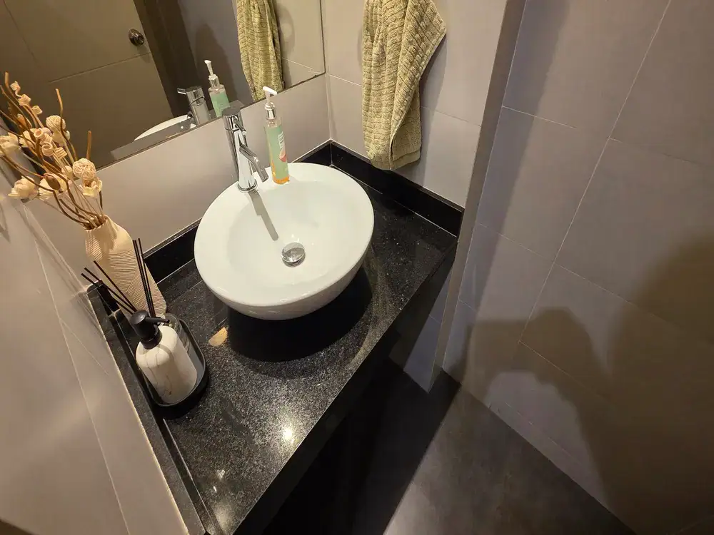 Oval white vessel sink on a black granite countertop with a chrome faucet, soap dispenser, and a beige towel hanging nearby, plus a vase of dried decorative stems in the corner.