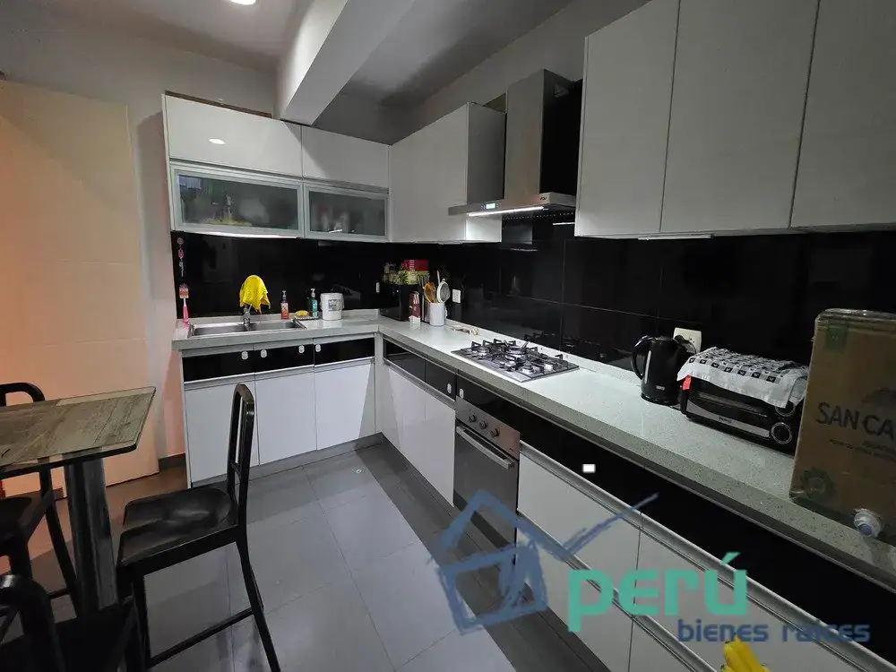 Modern L-shaped kitchen with white cabinets, black backsplash, and stainless appliances including gas stove and oven; sink area with drying rack and kettle nearby.
