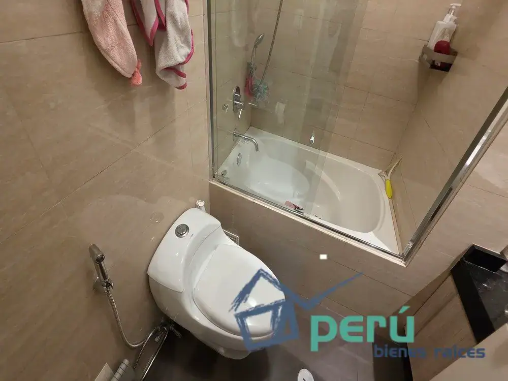 Bathroom with a toilet and glass-enclosed shower, towels hanging on the wall, and a logo watermark reading 'PERÚ bienes raíces'.