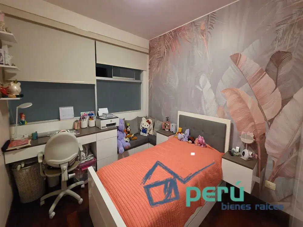Child's bedroom with a white bed and coral quilt, tropical leaf wallpaper, and a study desk along the left wall with a chair and shelves nearby. Plush toys sit on the bed and window blinds are above the desk area.