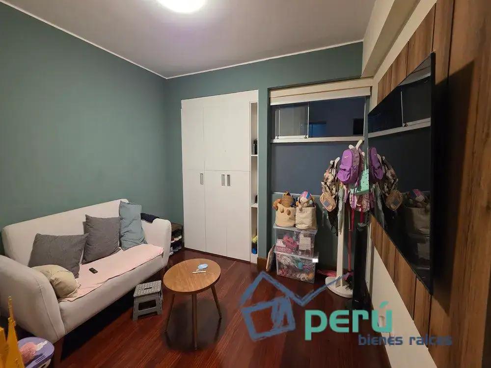 Cozy living room with a white sofa on teal walls, round coffee table, and a white storage cabinet. Footwear and baskets nearby suggest a family space.