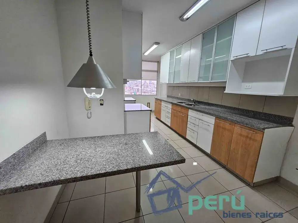 Modern L-shaped kitchen with granite countertops, white upper cabinets and wood lower cabinets; a metal pendant light hangs over a small island with a window showing a city view in the background.