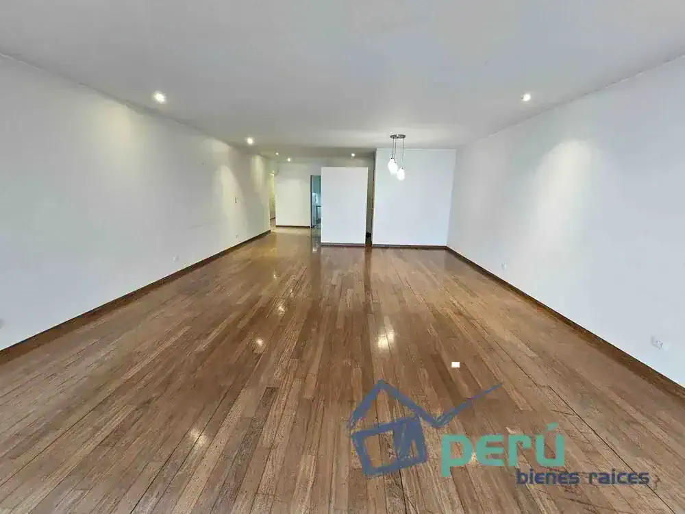 Spacious empty living room with hardwood floors, white walls, recessed lighting, and a doorway at the far end.