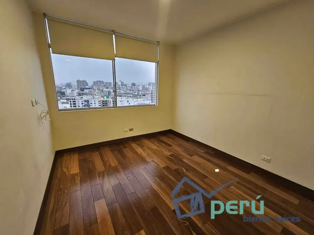 Empty room with dark wooden floor, beige walls, and a large window with blinds showing a city skyline.