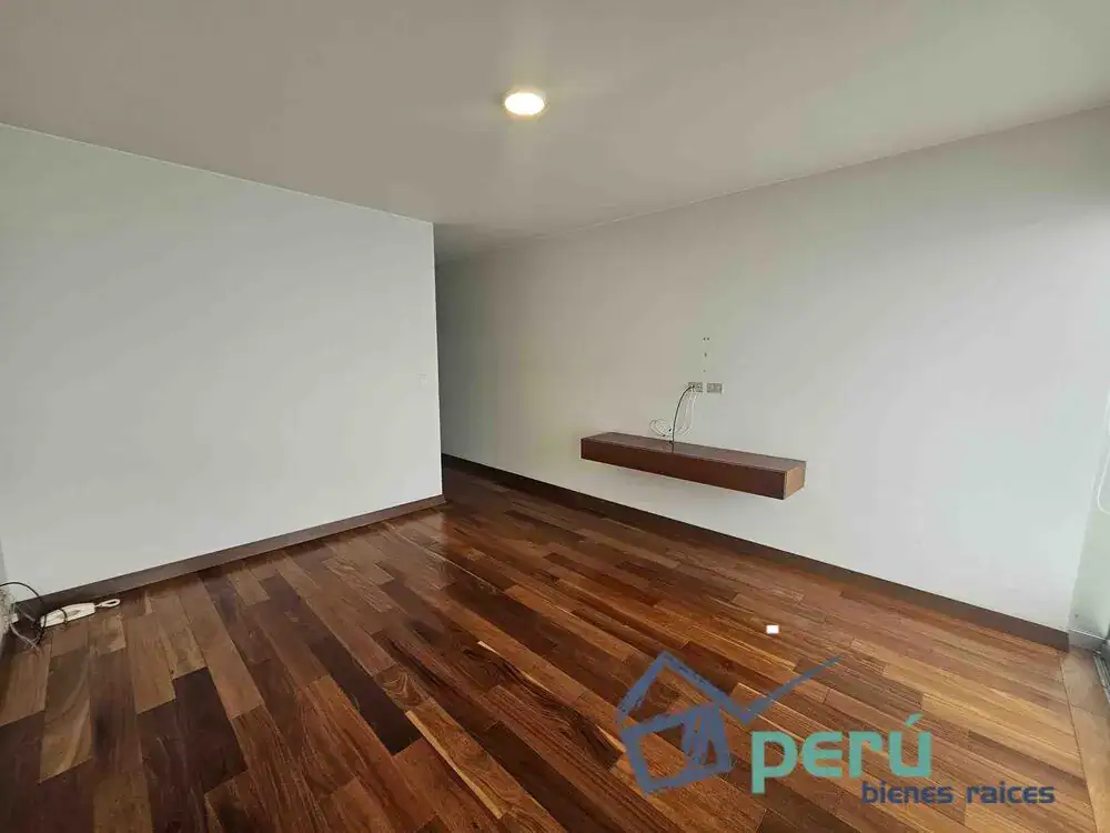 Empty living room with dark hardwood floor, white walls, and a floating wooden shelf on the right wall.