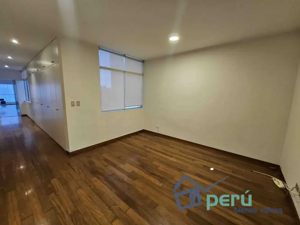 Empty unfurnished room with hardwood floors, white walls, and a large window; hallway with built-in cabinets on the left side visible in the background.