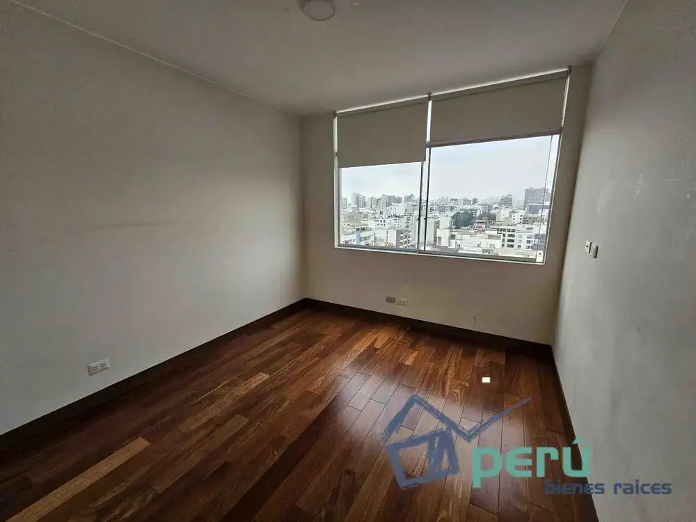 Empty room with dark wood flooring and a large window offering a city skyline view; blinds are partially drawn.