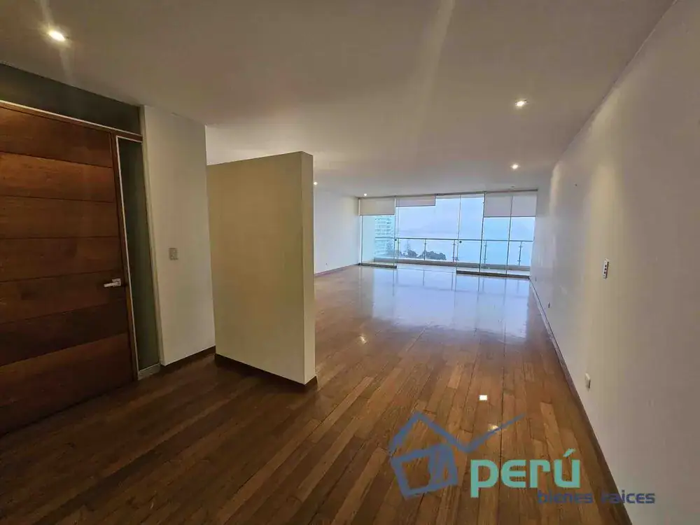 Spacious empty living area with wood floors and large glass doors overlooking the sea or city skyline, watermark at bottom corner.